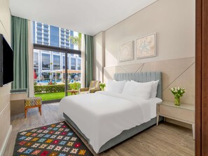 A modern hotel room with a large bed, a TV, a small table, and a view of the outdoor pool area. - Rixos Premium Alamein (El Alamein)