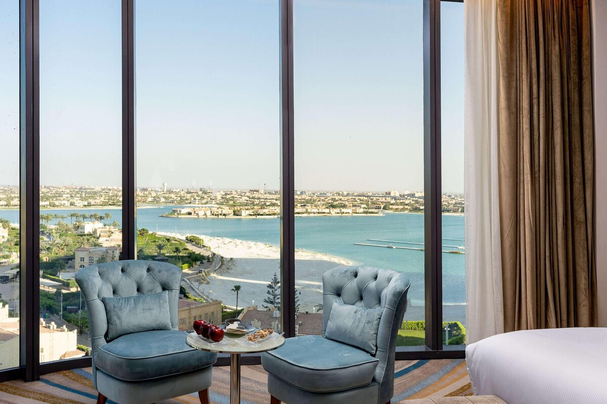 Une chambre d’hôtel dotée d’une grande fenêtre donnant sur une plage et sur le paysage urbain. On y trouve deux fauteuils rembourrés et une petite table ronde sur laquelle est posé un plateau de fruits.