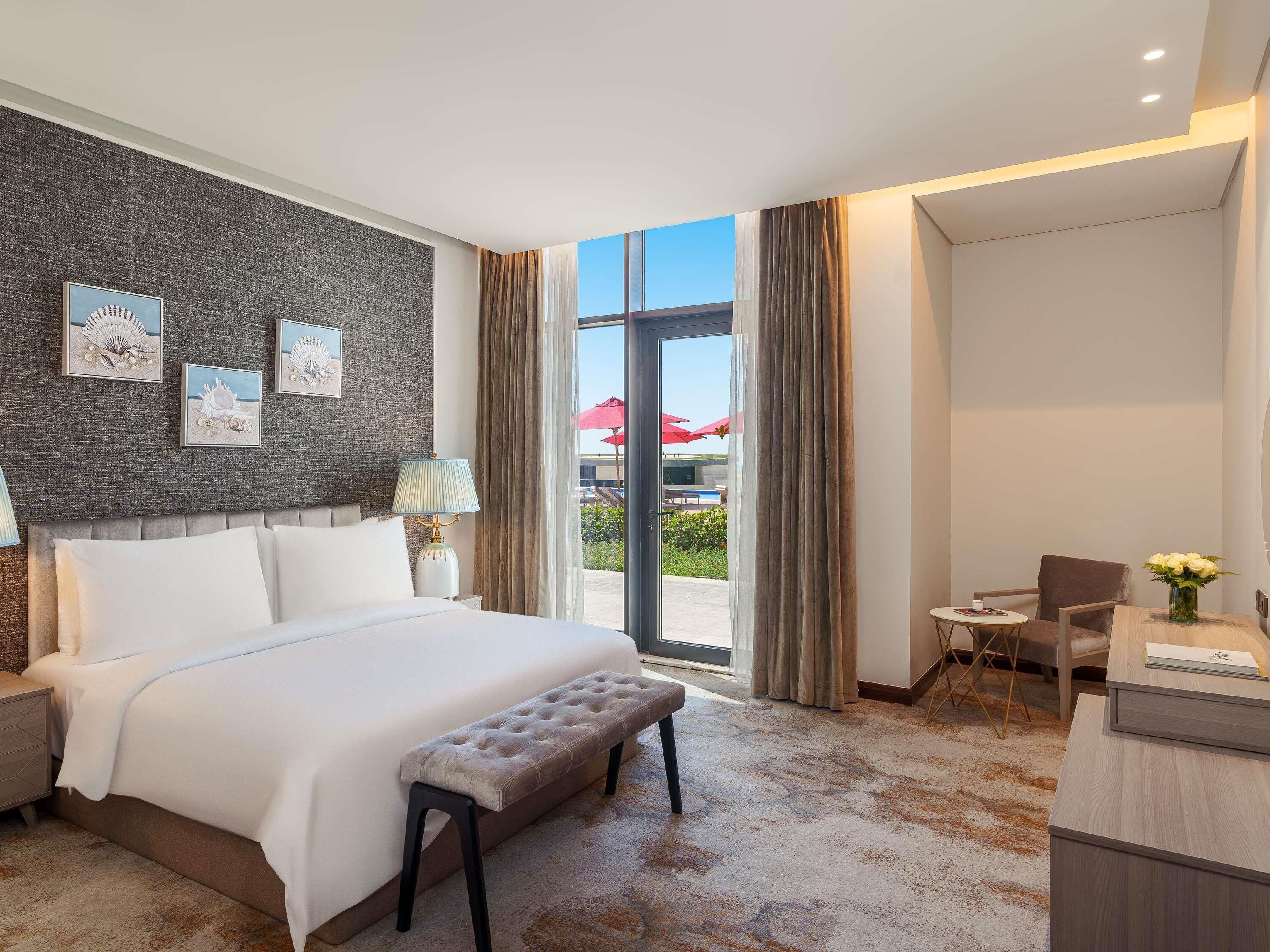 A hotel room with a large bed, a bench, a desk with a vase of flowers, and a view of the outdoor area.