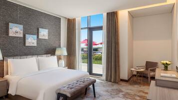 A hotel room with a large bed, a bench, a desk with a vase of flowers, and a view of the outdoor area.