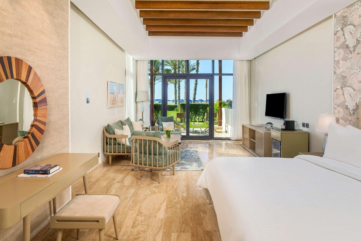 Ein modernes Hotelzimmer mit einem großen Bett, einem Schreibtisch, einem Sessel, einem Fernseher und einem Balkon mit Blick auf Palmen.