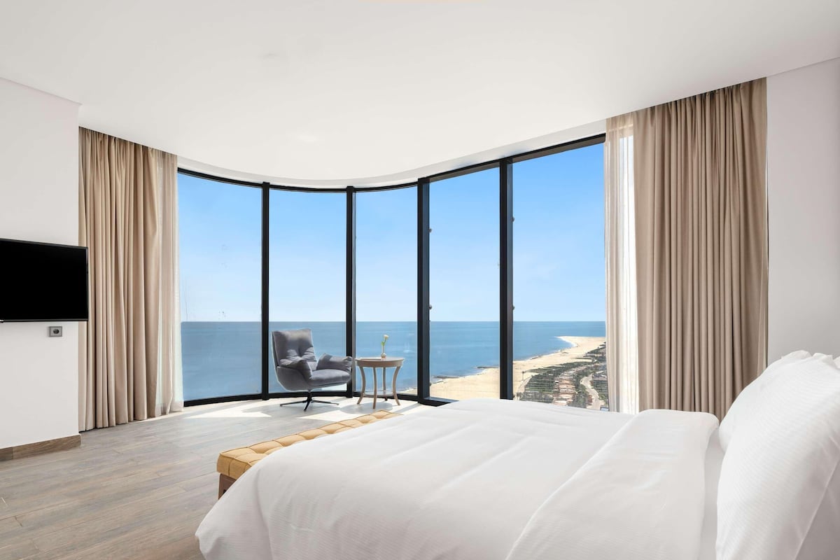 A bedroom with a large bed, a TV mounted on the wall, a small table with a vase, and a view of the beach through floor-to-ceiling windows.