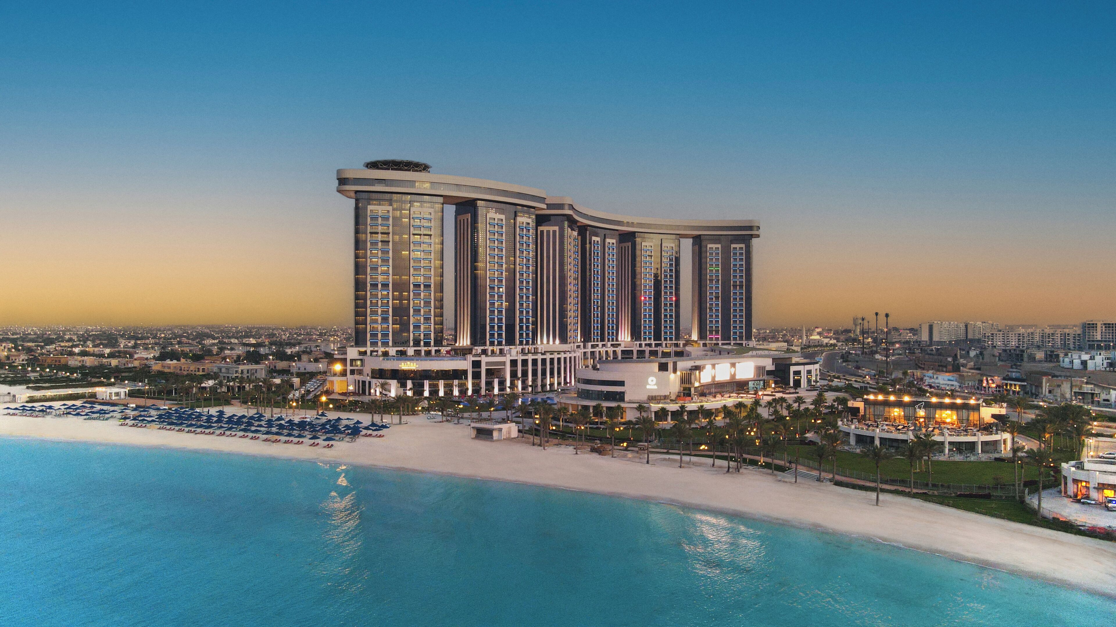 Un hôtel de grande hauteur situé en bord de mer, comprenant de nombreux étages, un espace piscine et une façade bien éclairée.