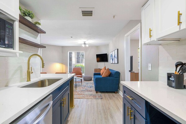 Una cocina moderna con encimeras blancas, armarios oscuros y tiradores dorados. Hay un ventanal con persianas, un sofá azul y un televisor montado en la pared.