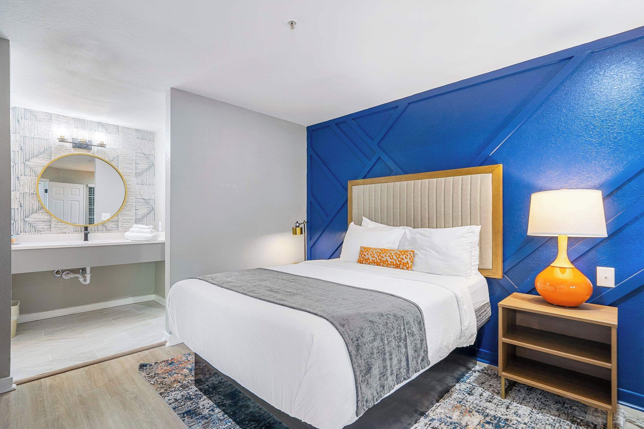 A modern hotel room with a large bed, a bedside table with a lamp, a bathroom with a round mirror, and a blue accent wall.