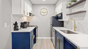 Une cuisine compacte avec des meubles blancs, un îlot central bleu, des appareils en acier inoxydable et une horloge dorée au mur.
