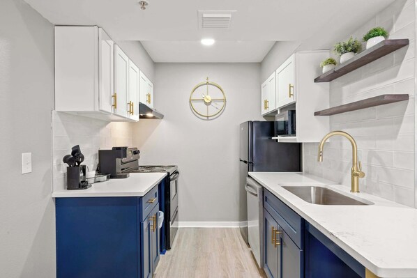 Una cocina compacta con armarios blancos, una isla azul, electrodomésticos de acero inoxidable y un reloj dorado en la pared.