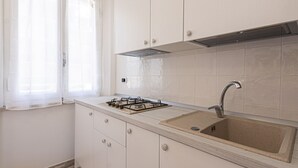 Una cocina con gabinetes blancos, una estufa a gas, un lavamanos de acero inoxidable y una ventana con cortinas traslúcidas.