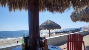 A beachside table with a thatched umbrella, bottles, and a red chair. - Condo Mirador with Rooftop Ramada Near Sea of Cortéz, El Malecon, and Manny's. (Puerto Peñasco)