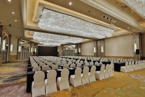 A conference room with rows of tables and chairs, large chandeliers, and a carpeted floor.