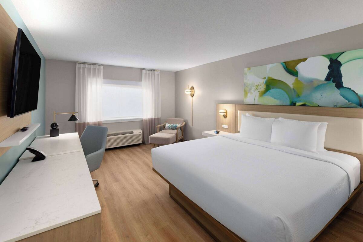 A hotel room with a bed, a desk, a chair, a flat-screen TV, a window with curtains, and a wall-mounted artwork.