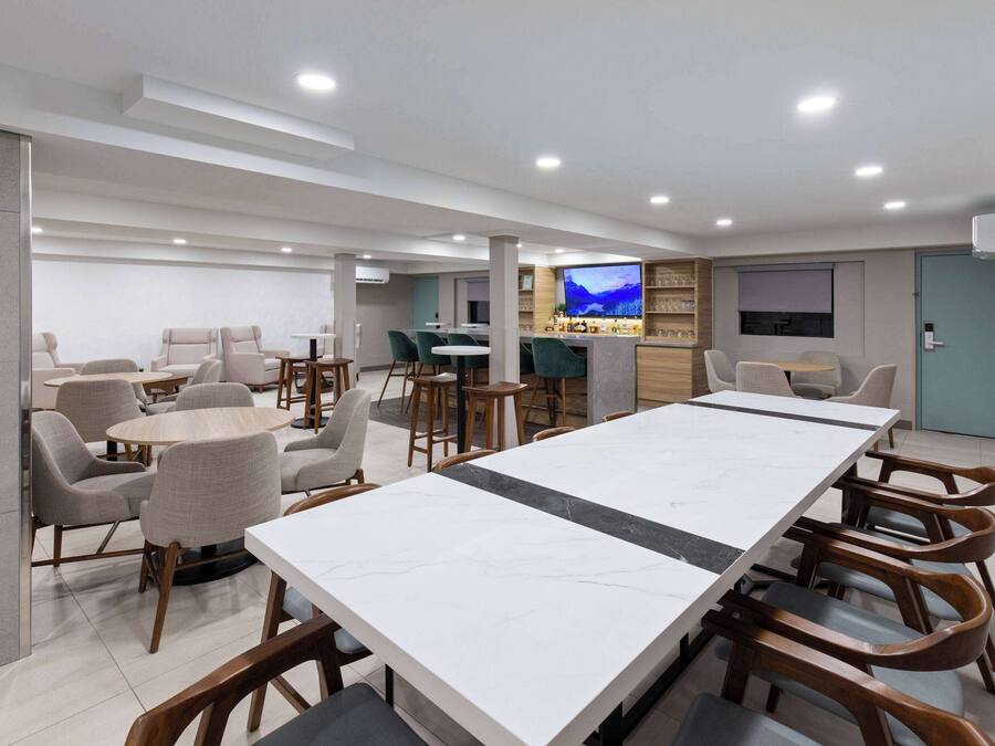 A modern hotel lobby with a long dining table, multiple seating areas, and a bar.