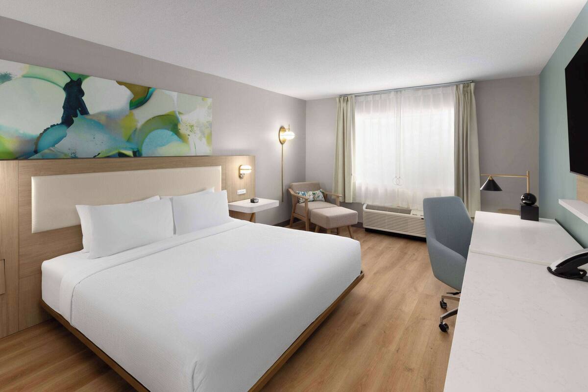 A hotel room with a large bed, a desk, a chair, a small table, and a wall-mounted TV.