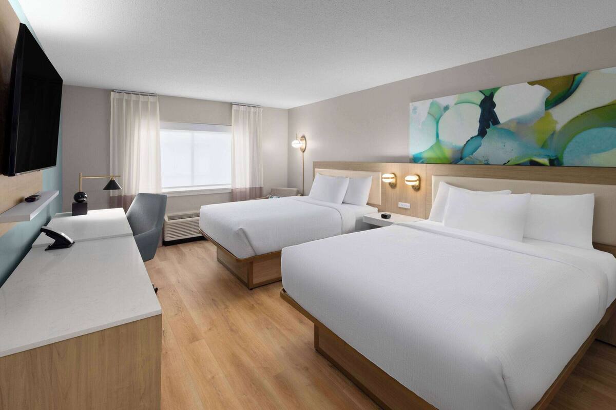 A hotel room with two beds, a desk, a chair, a TV, and a large abstract painting on the wall.
