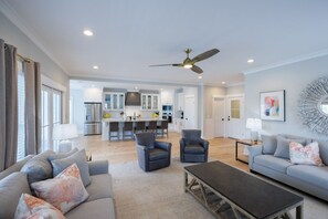 A spacious living room with a grey sofa, two armchairs, and a coffee table. A kitchen with white cabinets and a dining area with bar stools is visible in the background. - Tybee Ocean View & Private Pool - 1B Oceanview Court (Tybee Island)