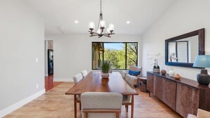 A spacious dining area with a long wooden table, chairs, and a chandelier. There is a large window providing a view of greenery outside. - Summit Lodge- Sleeps 10, 4 Full Bath, Pet Friendly (Prescott)