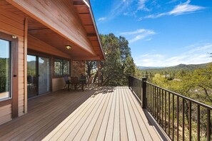 Um deck de madeira com guarda-corpo, uma mesa e cadeiras, e vista para uma paisagem arborizada.