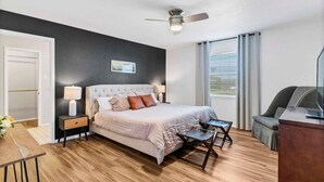 A bedroom with a bed, bedside table, lamp, chair, desk, and a ceiling fan.
