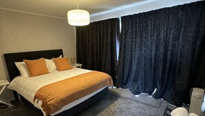A hotel room with a bed, a bedside table, a lamp, a window with curtains, and a radiator.