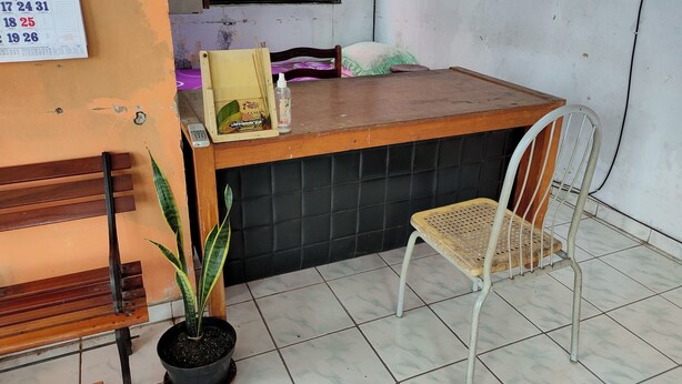 A reception area with a wooden counter, a white metal chair, a potted plant, and a bulletin board with numbers. - Homestay Wijaya Mulya By Zuzu (Borobudur)