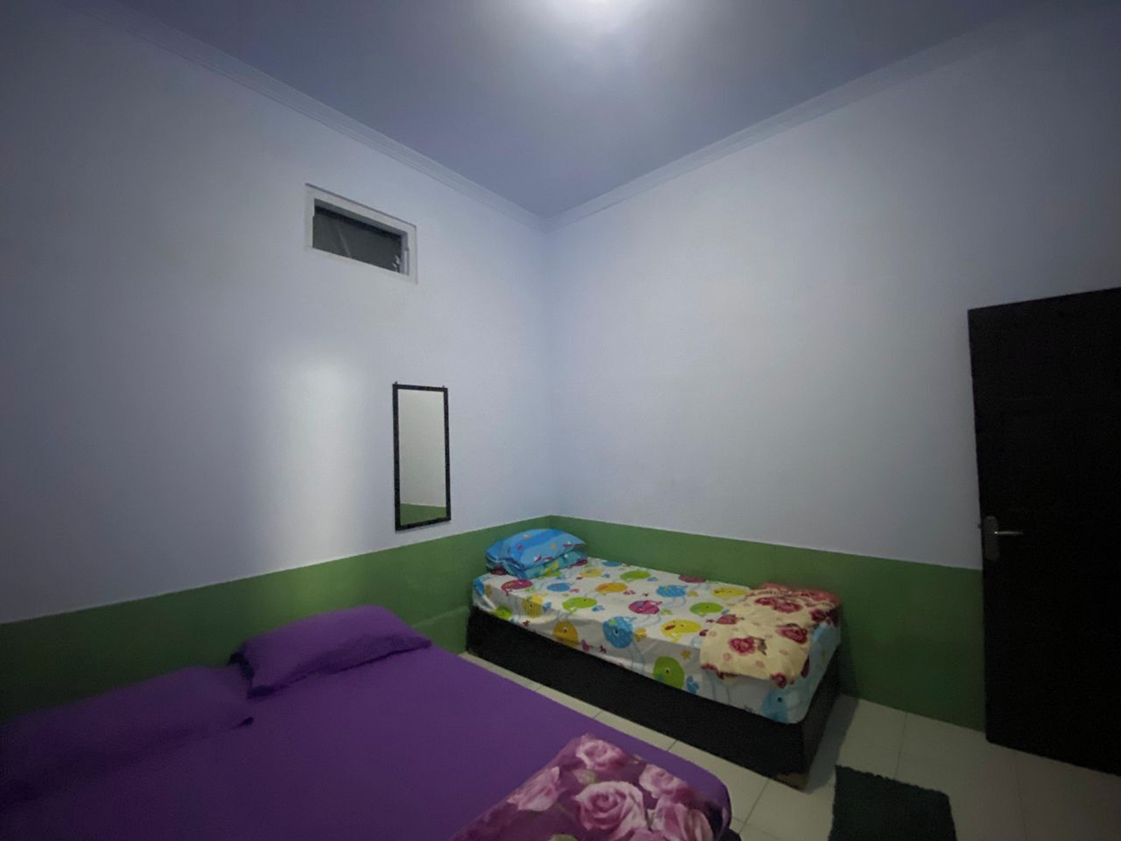 Ein kleines Zimmer mit einem Einzelbett, einer violetten Tagesdecke, einer bunten Bettdecke, einem kleinen Fenster und einer Tür.