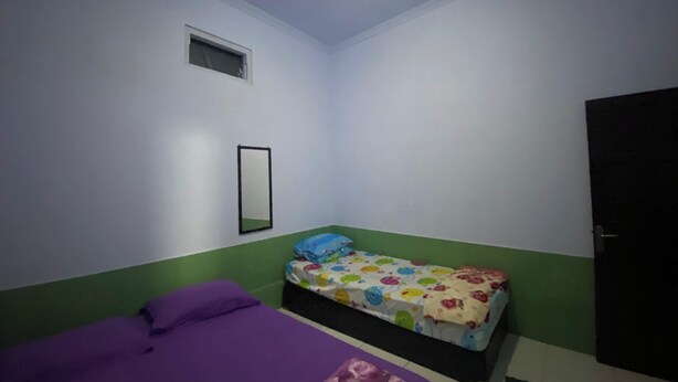 Una habitación pequeña con una cama individual, colcha morada, colcha colorida, una ventana pequeña y una puerta.