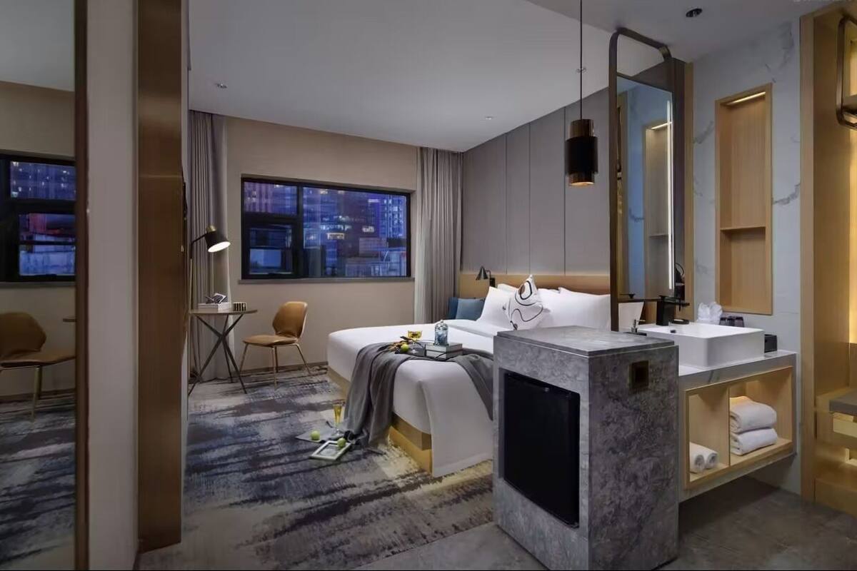 Una moderna camera d'albergo con un letto grande, una scrivania con una sedia, un'ampia finestra vista città, un bagno con un lavabo e uno specchio, e un grande specchio sopra il letto.