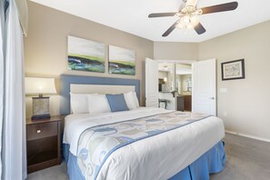 Une chambre à coucher comprenant un lit, une table de chevet, un ventilateur de plafond, des œuvres d’art et une porte ouverte donnant sur une salle de bain.