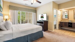 Une chambre d’hôtel avec un grand lit, un ventilateur de plafond, une télévision, un balcon donnant sur un espace verdoyant, et une salle de bain avec un miroir et un lavabo.