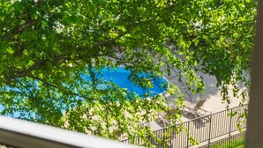 Une piscine entourée d’arbres verdoyants et d’un bâtiment à la façade blanche et au garde-corps noir.