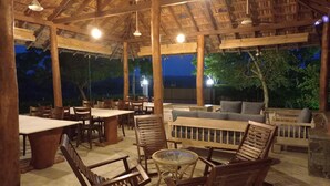 An outdoor dining area with wooden furniture and a thatched roof. Tables and chairs are arranged for dining, and there is a seating area with cushions. - yala leopard lodge (Kirinda)