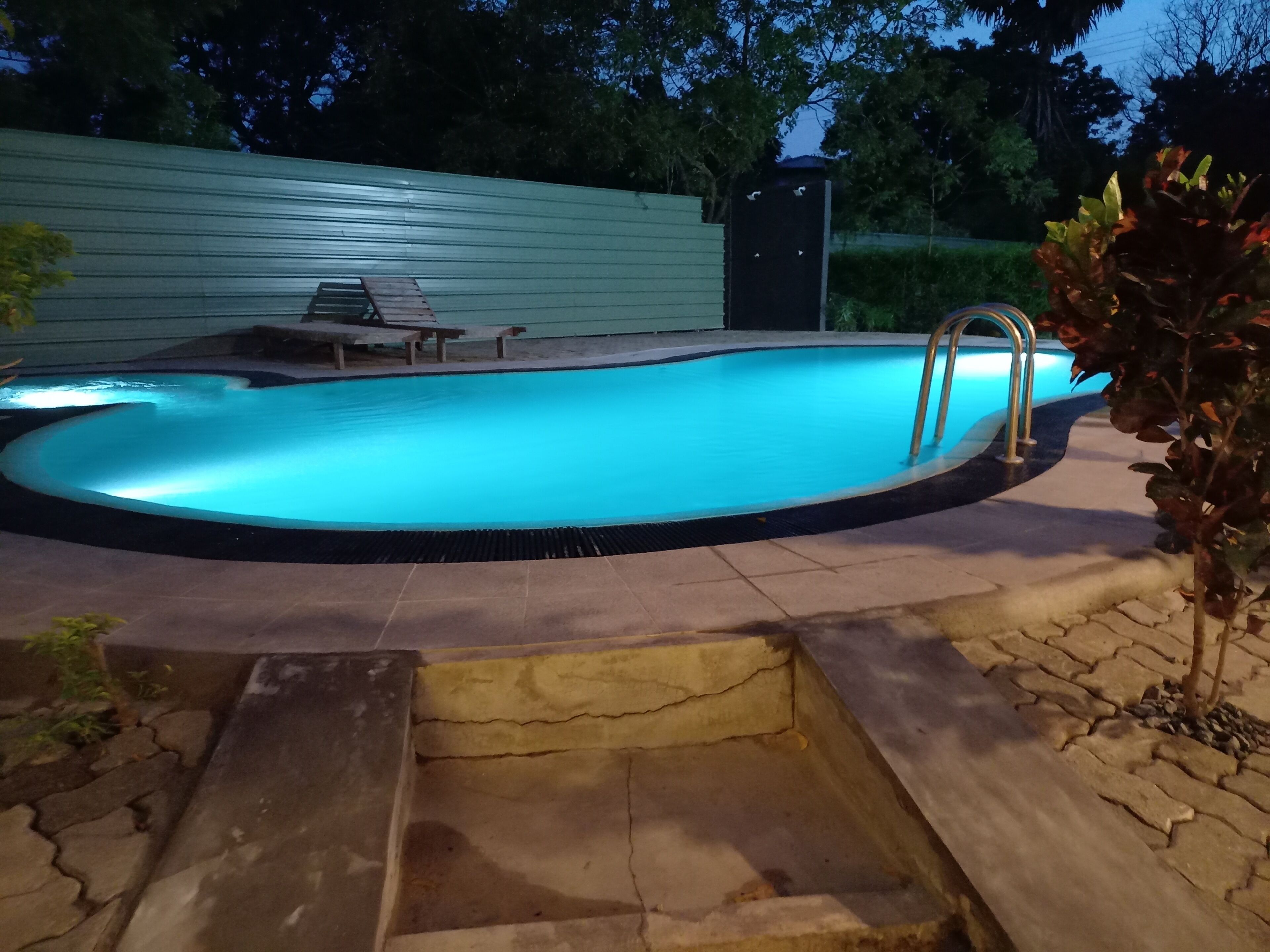 A swimming pool with a metal ladder, surrounded by a stone deck and a wooden deck with lounge chairs.