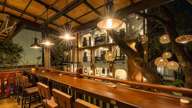 A bar area with wooden chairs and tables, hanging pendant lights, and a large tree in the background. - Shamrock Chiangmai Hotel - Adults Only (Chiang Mai)