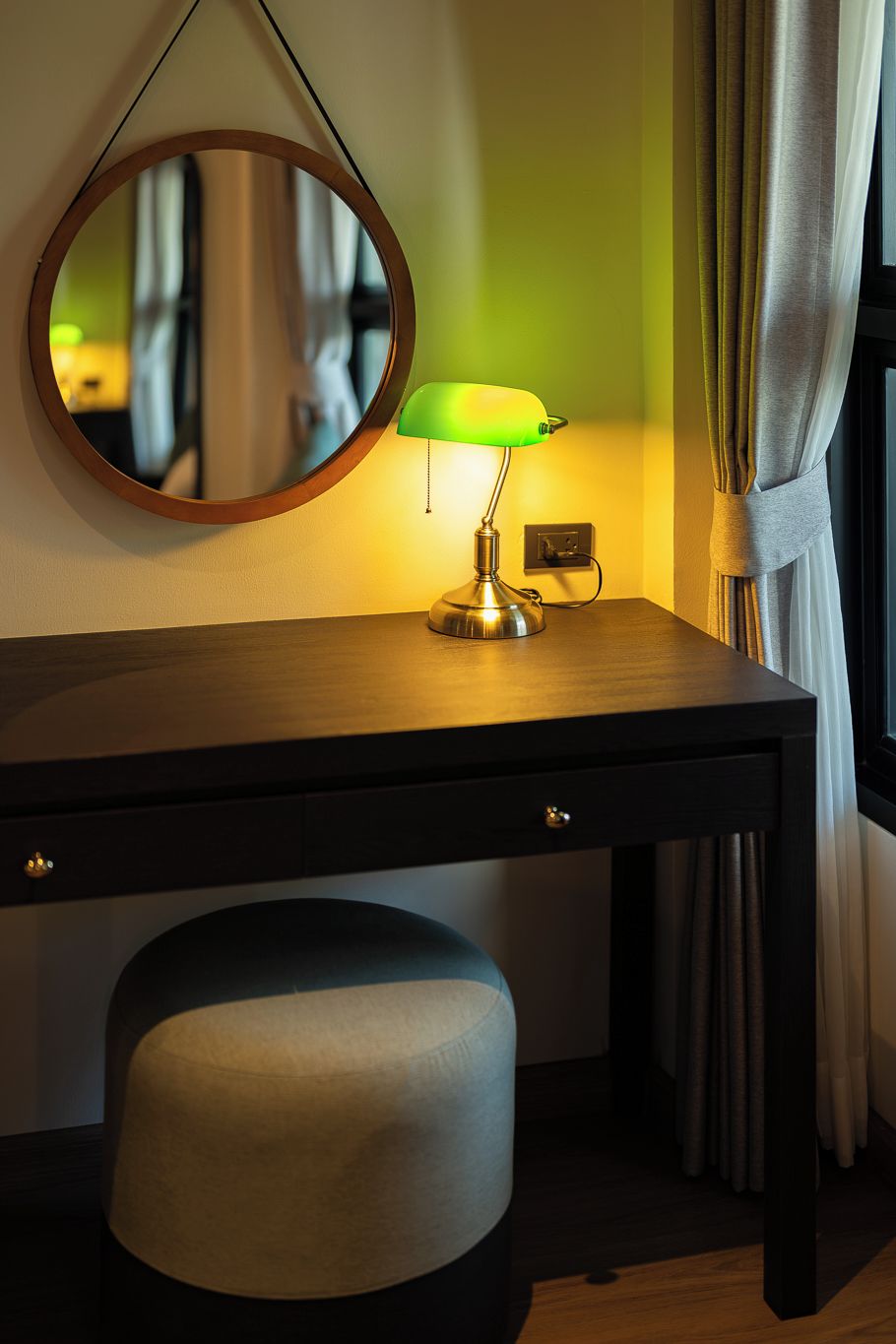 A wooden desk with a green desk lamp, a round mirror, and a cushioned stool.