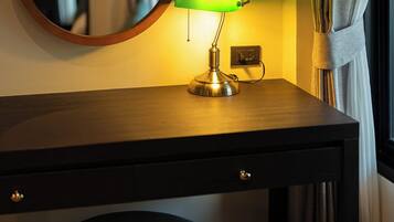 A wooden desk with a green desk lamp, a round mirror, and a cushioned stool.