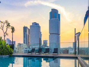A modern hotel with a pool, lounge chairs, and a view of tall buildings against a sunset sky.