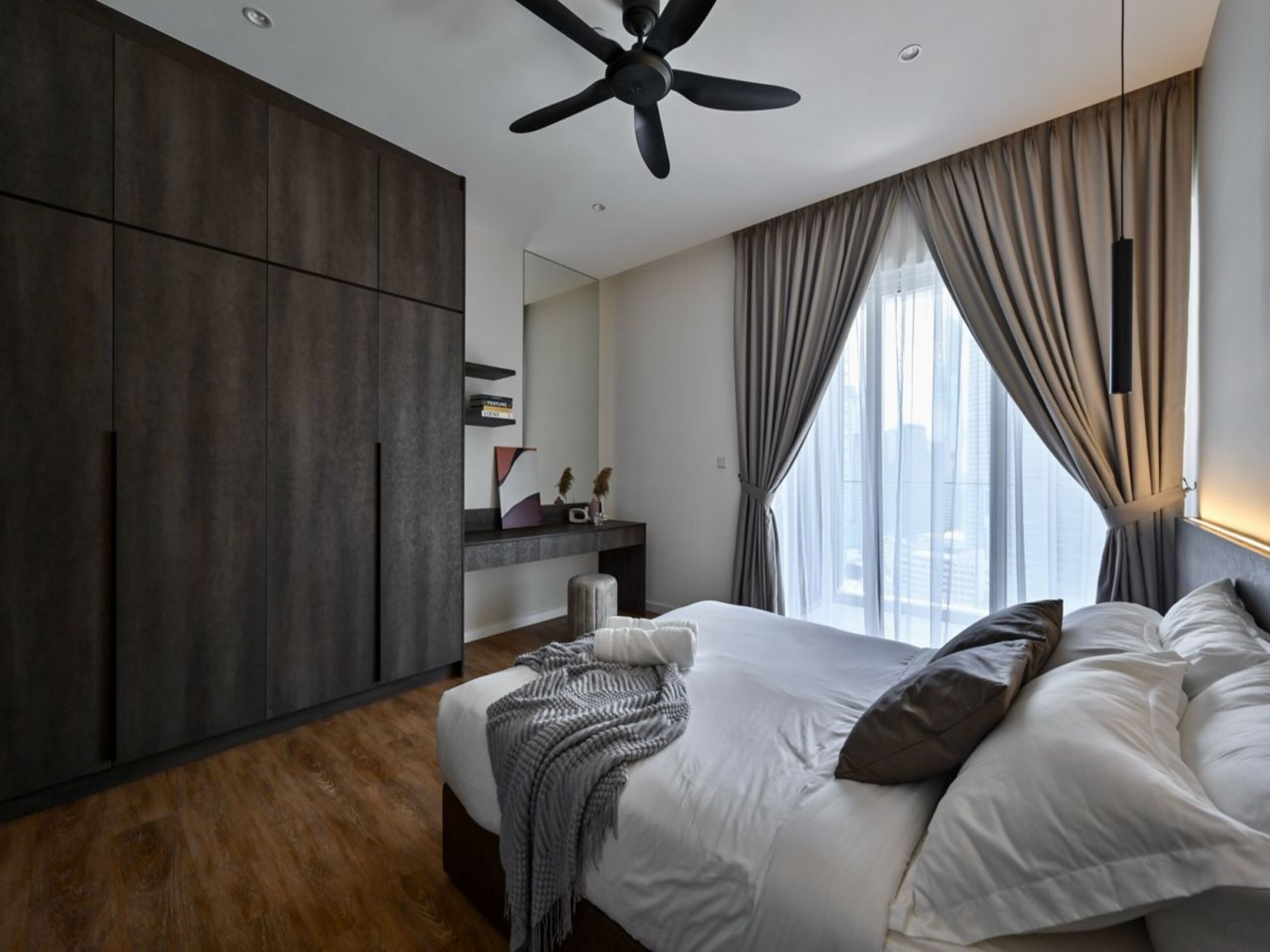 A modern hotel room with a large bed, dark wooden wardrobe, a ceiling fan, and a city view through large windows.
