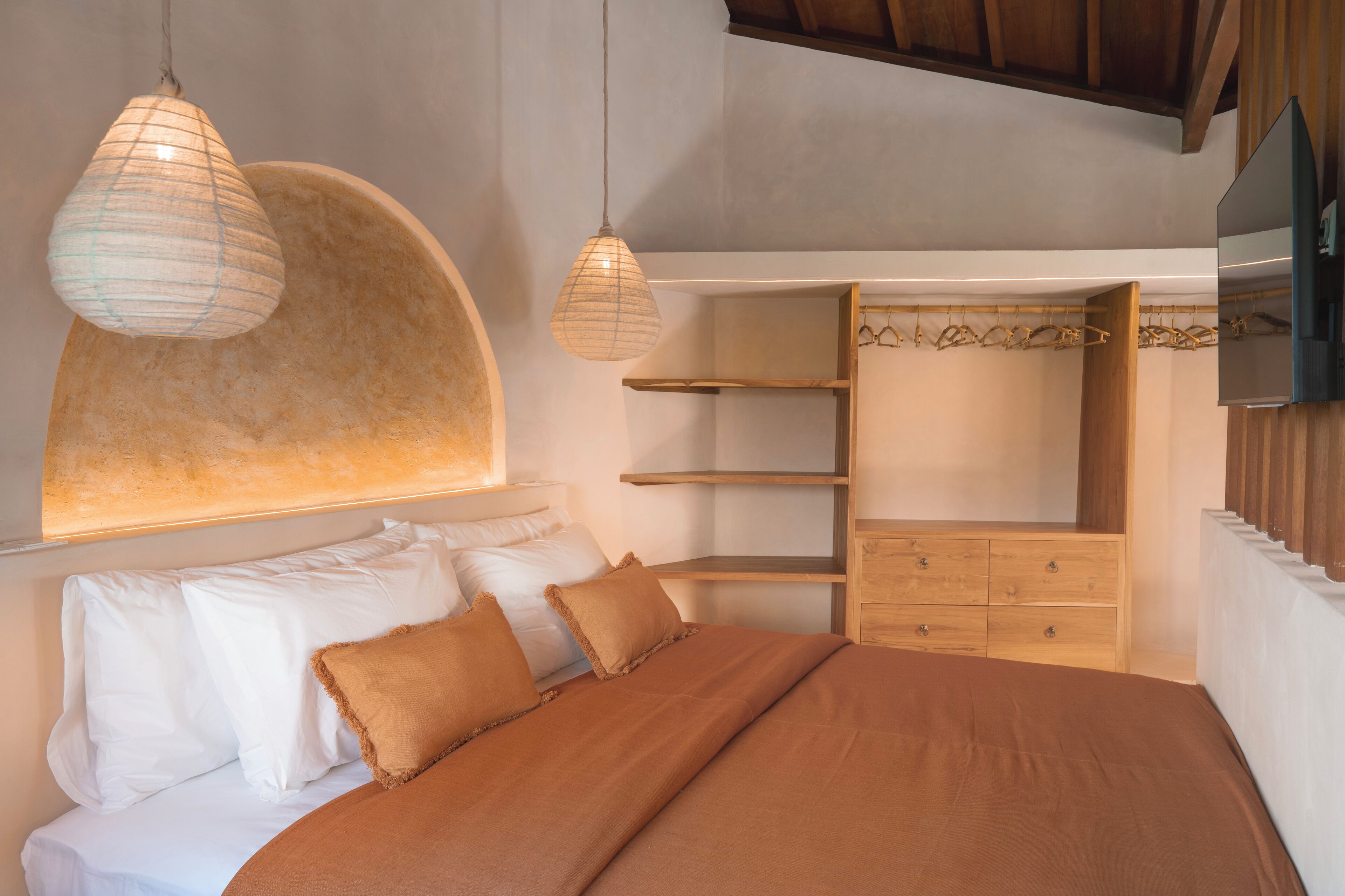 A bedroom with a bed, white pillows, a brown blanket, wooden shelving, and hanging clothes.