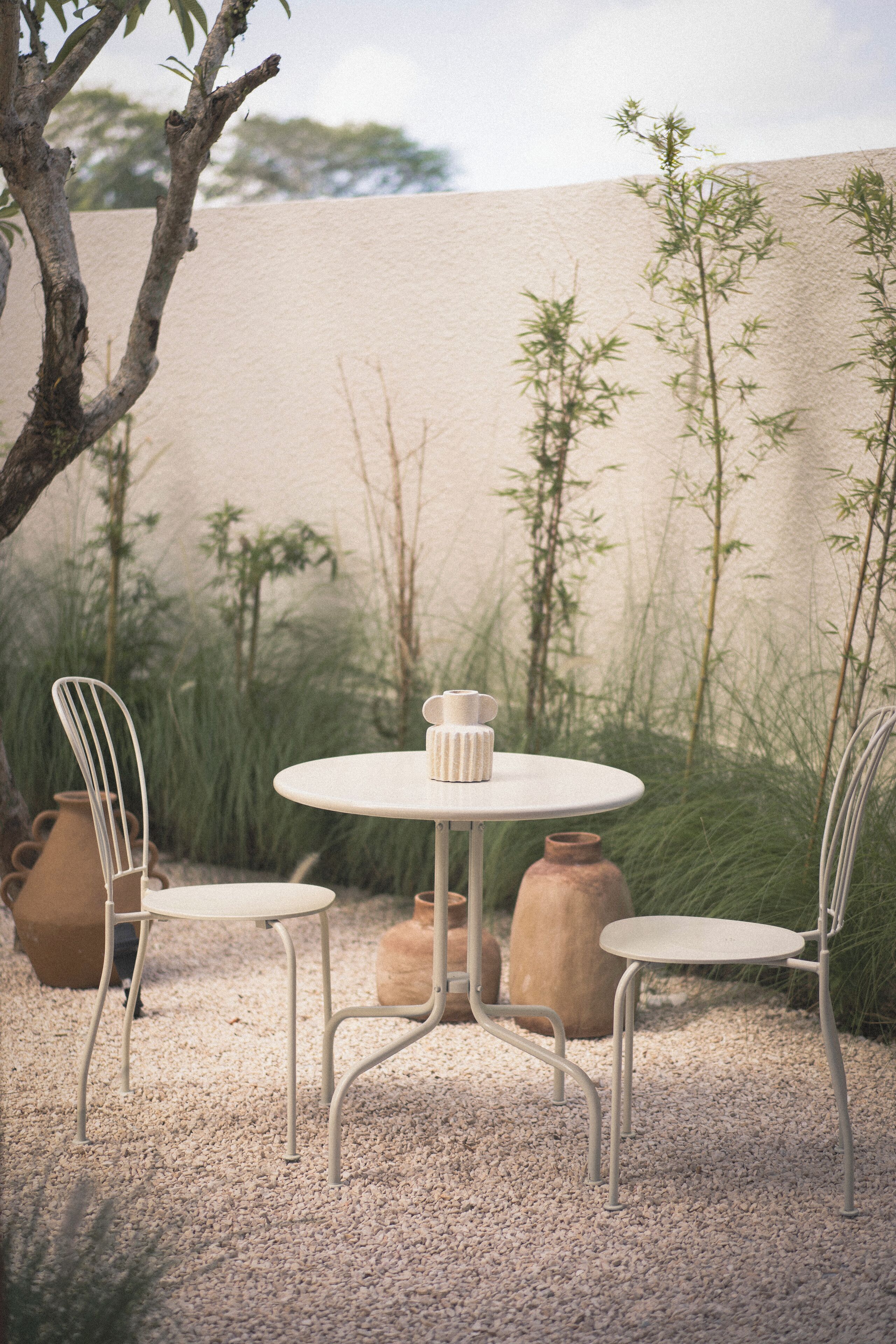 A round table with two chairs, surrounded by gravel and plants, with a beige wall in the background.