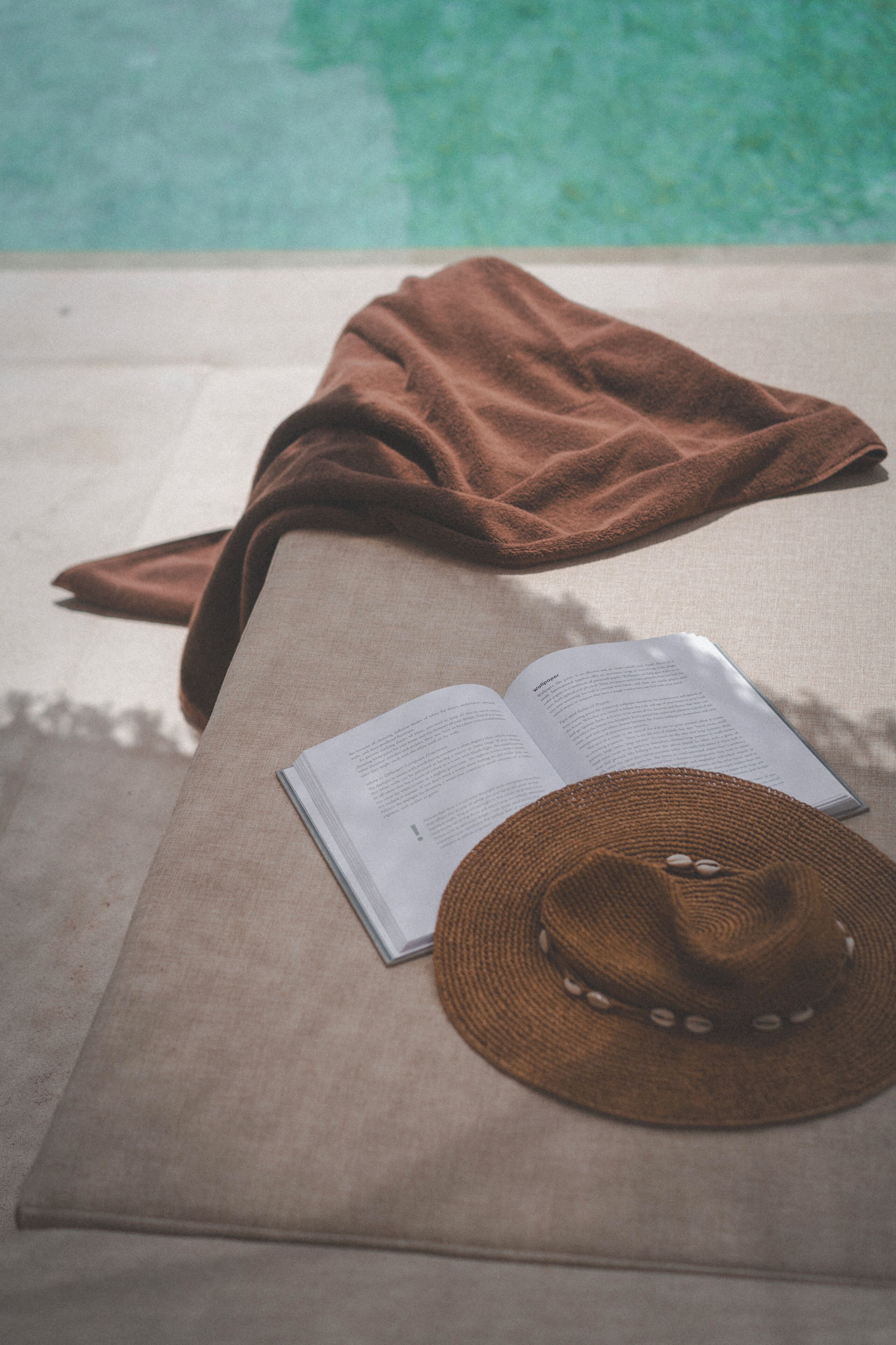 Ein Handtuch, ein Buch und ein Hut liegen auf einem beigen Sofa am Pool.