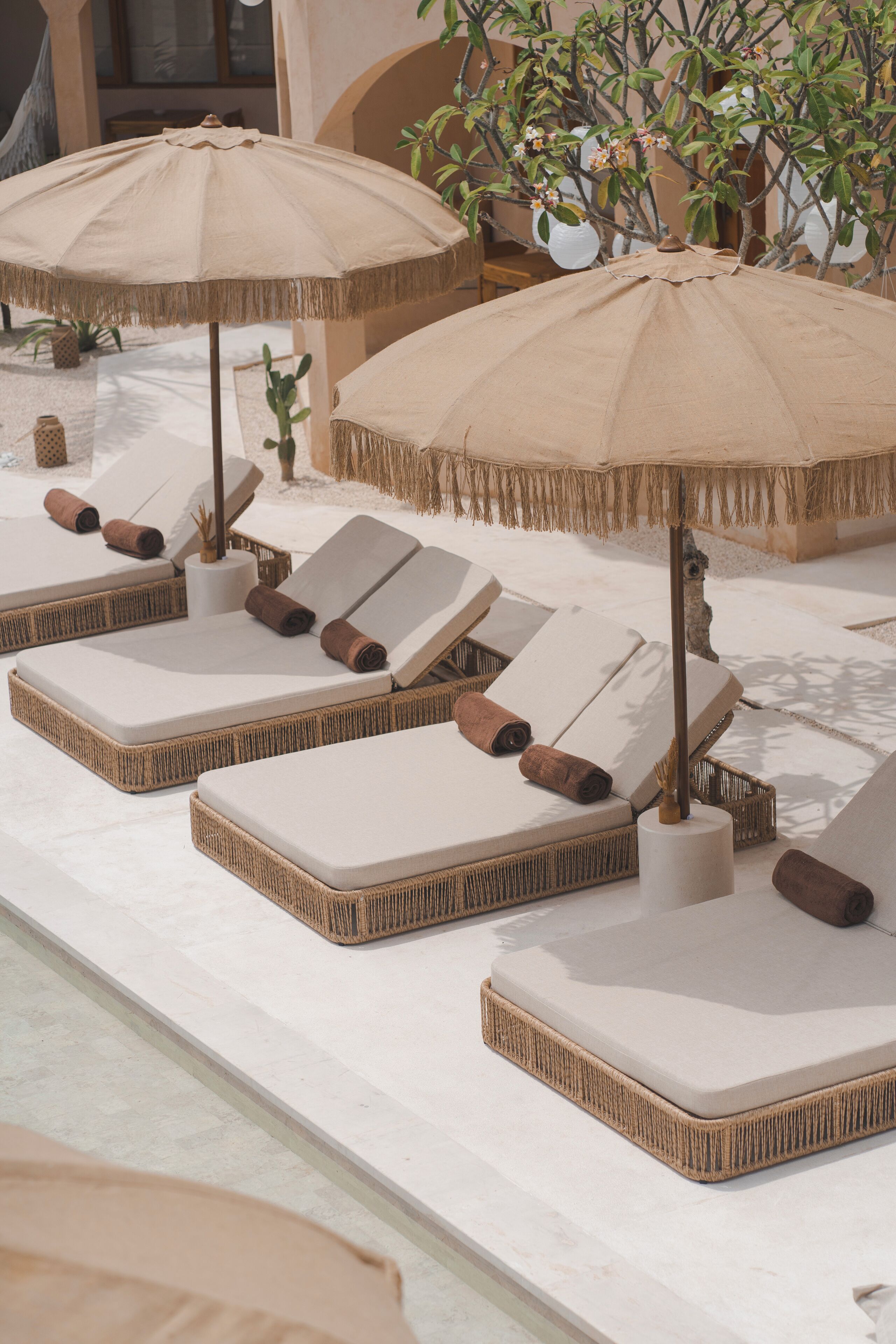 A row of sun loungers with beige cushions and straw umbrellas, arranged along a poolside.