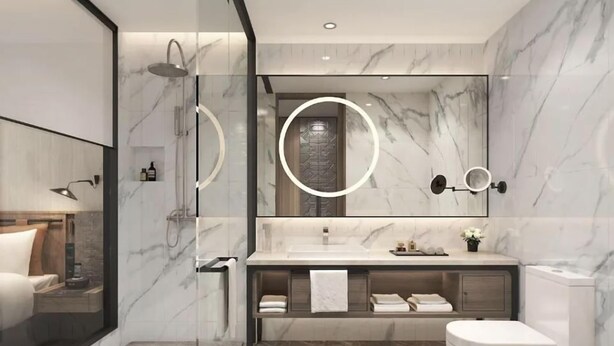 Un baño moderno con cabina de ducha de vidrio, suelo de baldosas de mármol, un espejo grande con iluminación circular y un inodoro blanco.