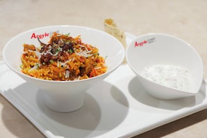 Una bandeja blanca con dos recipientes de comida: uno con un plato de arroz, verduras y carne, y el otro, una crema blanca.