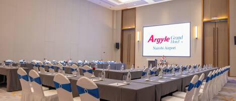 A conference room with a long table, chairs with blue sashes, and a projector screen displaying 'Argyle Grand Hotel Nairobi Airport'.