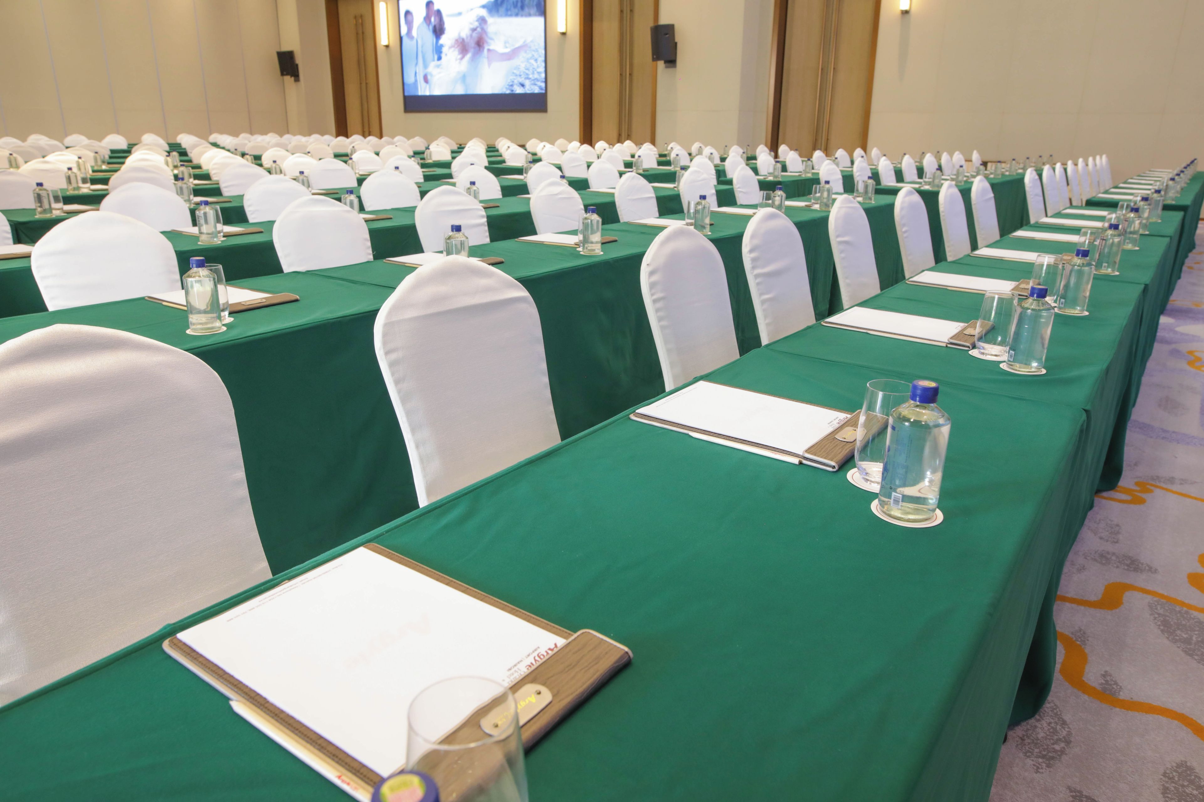 A conference room with rows of tables, each with a green tablecloth, white chairs, and water bottles.