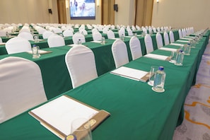 Een vergaderzaal met rijen tafels, elk met een groene tafelkleed, witte stoelen en waterflesjes.