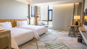 A hotel room with two beds, a small table with a lamp, a chair, and a view of the outdoors.