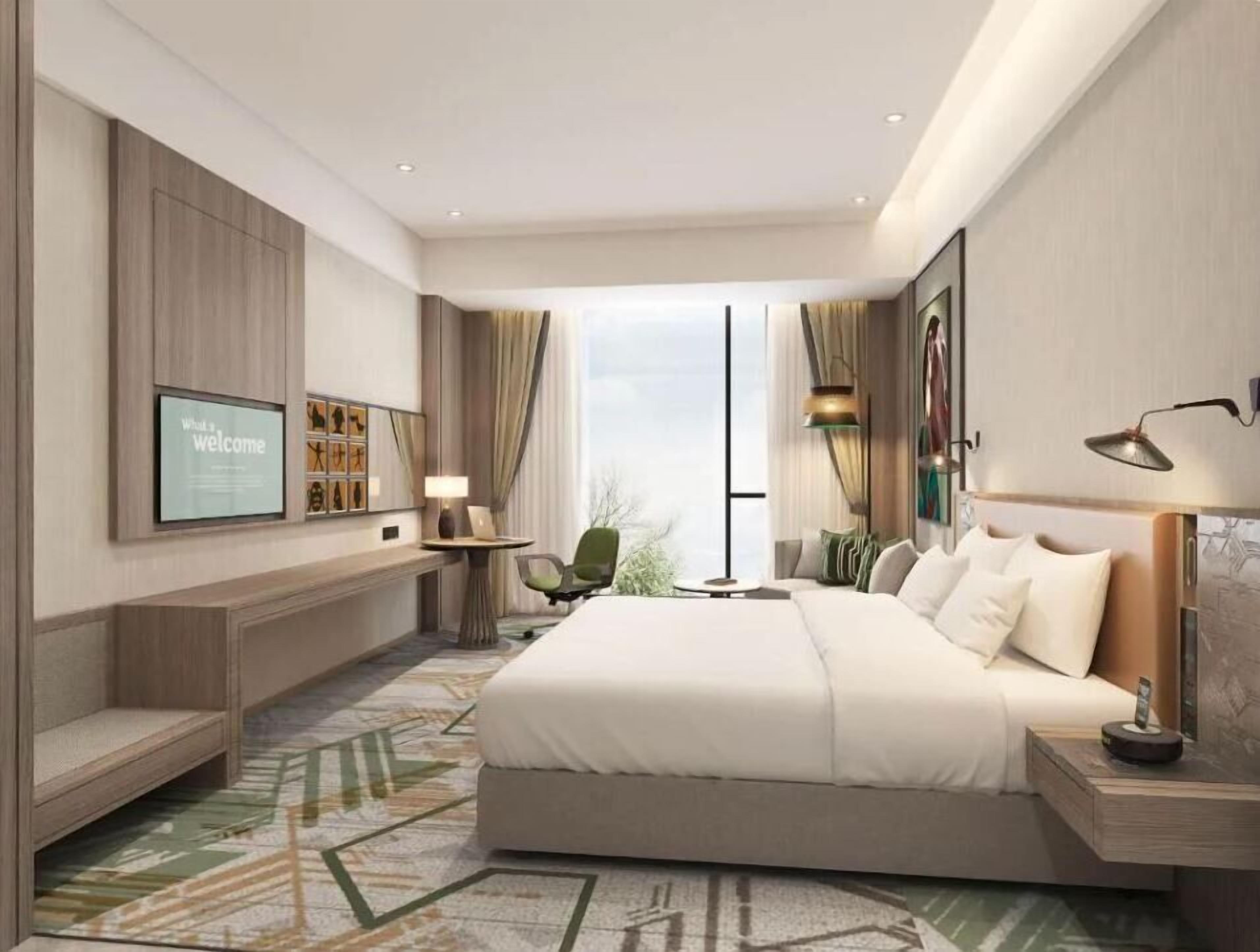 Quarto de hotel moderno com uma cama grande, televisão na parede, escrivaninha com luminária e vista para o exterior.