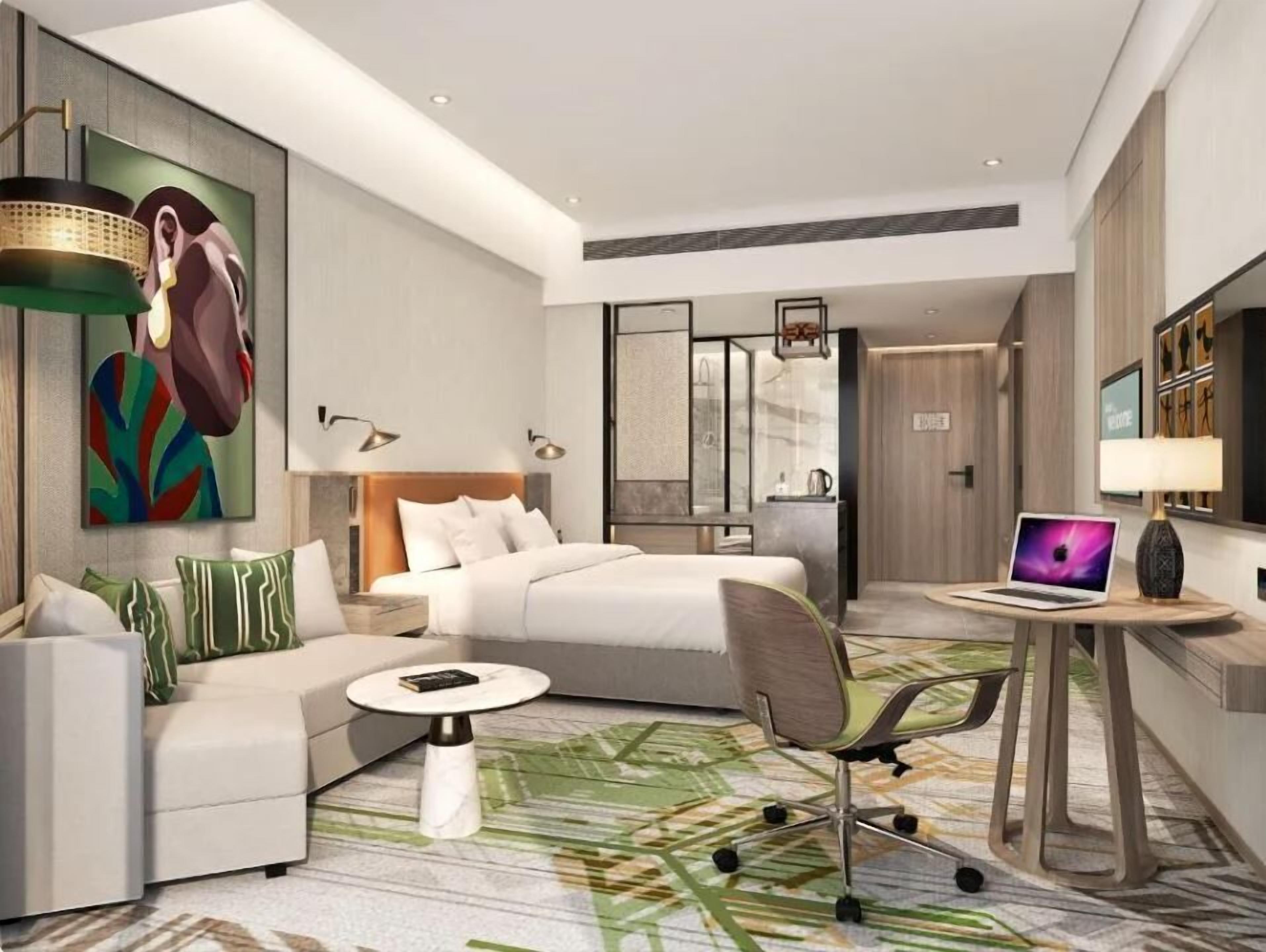 Quarto de hotel moderno com cama, sofá, escrivaninha com um laptop e um quadro na parede.