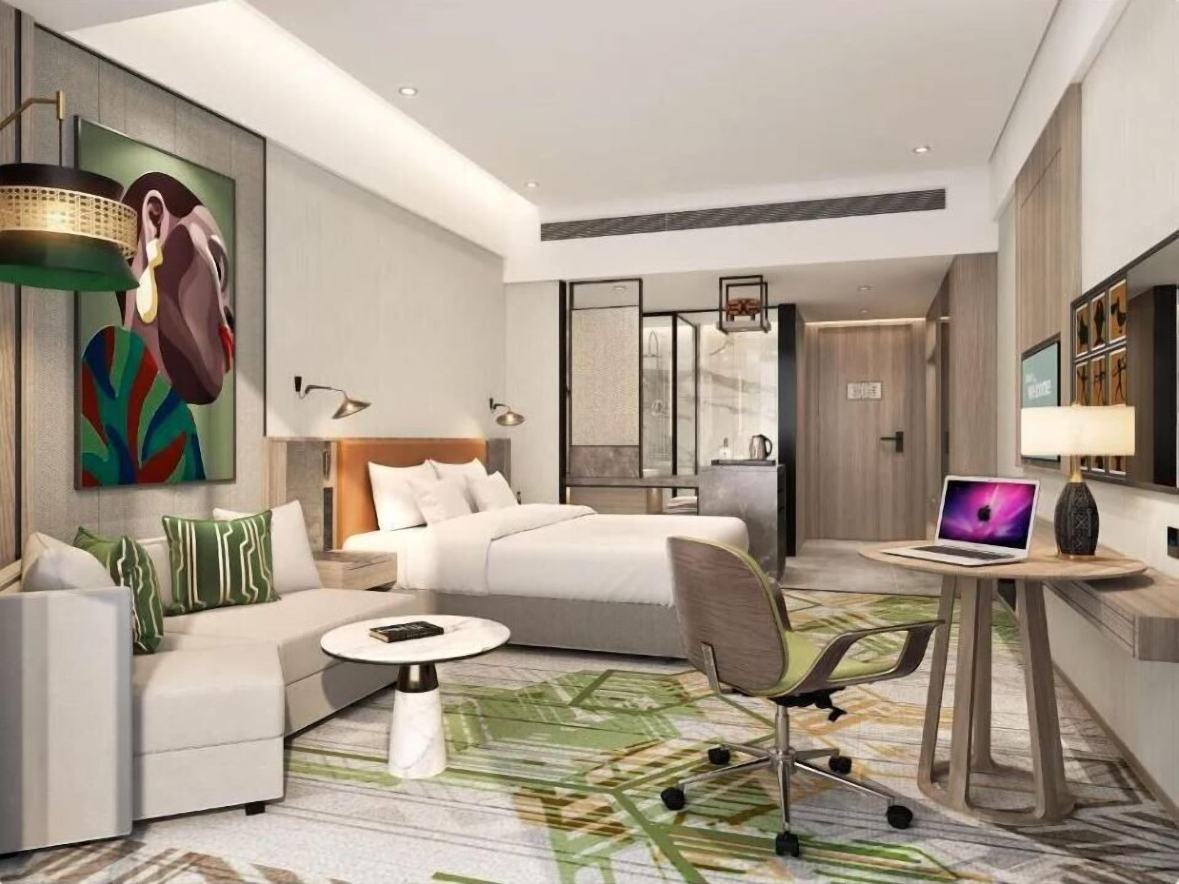 Quarto de hotel moderno com cama, sofá, escrivaninha com um laptop e uma pintura grande na parede.